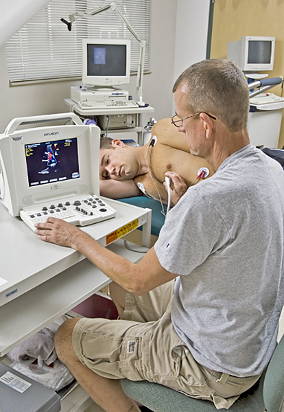 Shirtless man lays on side with nodes attached to his chest. Seated man holds a meter up to the shirtless man's chest while staring at a digital interface of the information gathered.