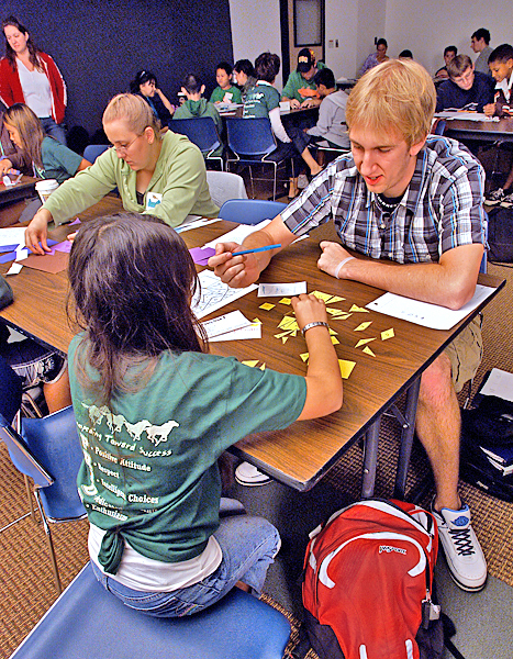 College-aged male sits at the end of a table across from a young girl playing a game with various rectilinear shapes cut out of yellow paper scattered between them on the table.