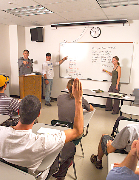In a class setting, a group of three people lead a discussion, with two people pointing to a white board and one man behind a podium pointing at a man in a desk who is raising his hand.