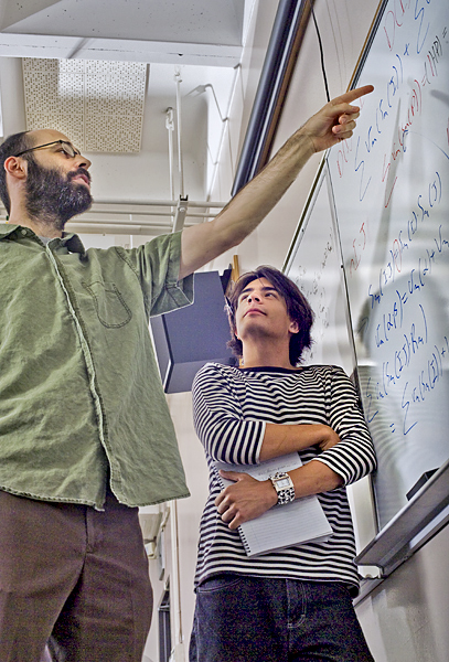 Man in green shirt stands and points at a mathematical equation written on a white board while a man in a black and white striped shirt leans against the board holding a note book.
