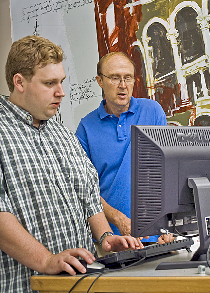 Two men stand at a computer. One man types and the other man is speaking.