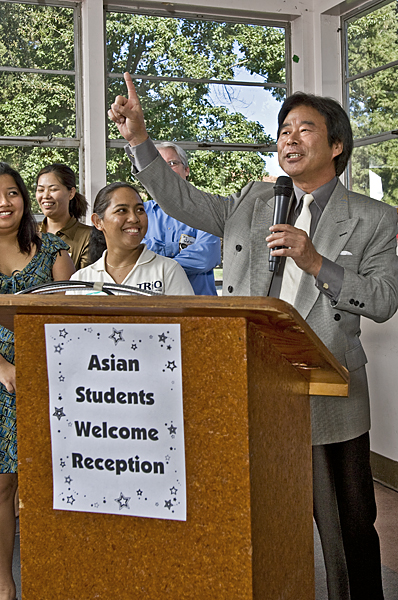 Man in gray suit holding a microphone points up into the air from behind a podium with a poster on it front reading "Asian Students Welcome Reception."
