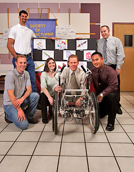Four men and one woman gather around a display and a manufactured prototype of a wheelchair created by engineering students.