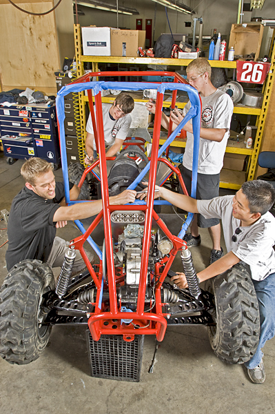 Four men stand or kneel around the four corners of a mechanized vehicle. One man in front passes another man a socket wrench. The open frame of the vehicle still has protective plastic covering parts.