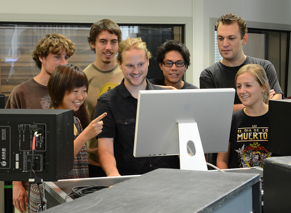 A diverse group of 6 students surround one man wearing a black shirt who is smiling and standing at a computer monitor.  A woman standing to the left of the photo smiles and points at the monitor.