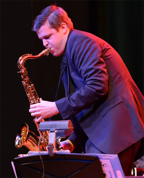 A man dressed in a dark colored suit, faces left and crouches playing the saxophone against a black background. A pink light cast from the musician's back overshadows a soft blueish light illuminating his front. In the foreground stands the top of a music stand with a head-lamp attached to the top.