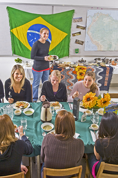 A Brazilian flag hangs over the service table for a classroom feast as a group of women sit on both sides of the dining table preparing to dig in.