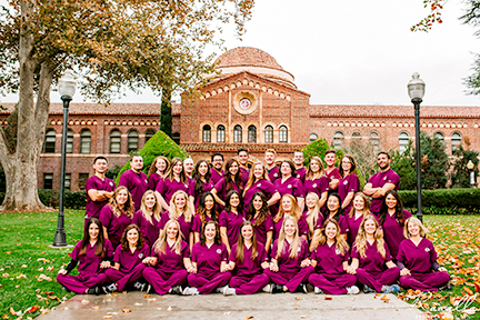 A group of students, men and women, wearing cardinal colored nurses scrubs pose together in front of Kendall Hall on the Chico State campus.