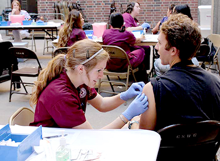 Woman, wearing cardinal colored scrubs, and light blue latex gloves prepares the arm of a young man for a flu shot to be administered.