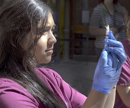 One woman, on the left of the photo, looks intently at a syringe, that she holds up while wearing blue latex gloves.