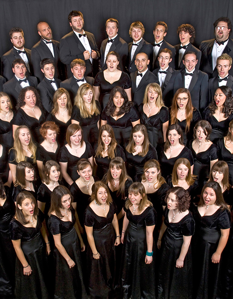 Six rows of students formally dressed sing in unison; some look at the camera and smile.
