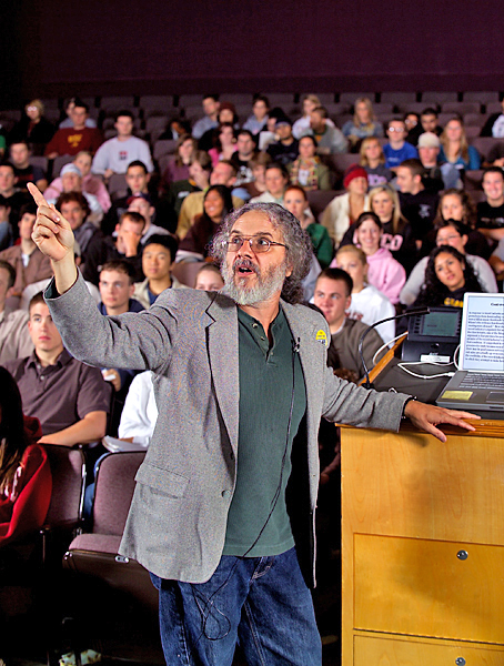 Man wearing glasses and a grey blazer opens out towards the camera raising his arm, pointing to direct the attention of the students sitting in the theater-style classroom.
