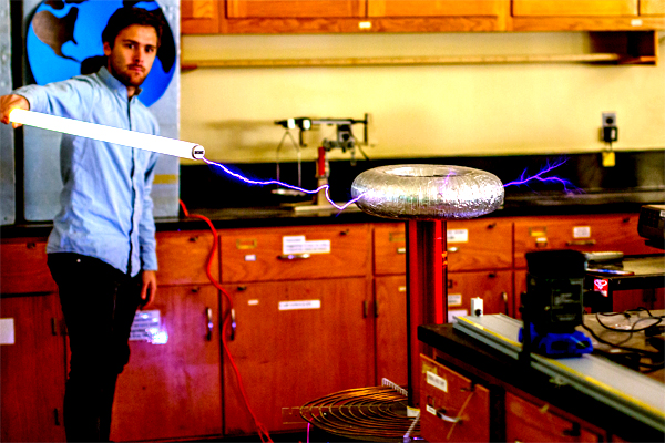 A man in light blue shirt and black pants, in what appears to be a science lab, points an incandescent light bulb towards a donut-shaped device wrapped in foil.  A light purple bolt of electricity connects the two objects.
