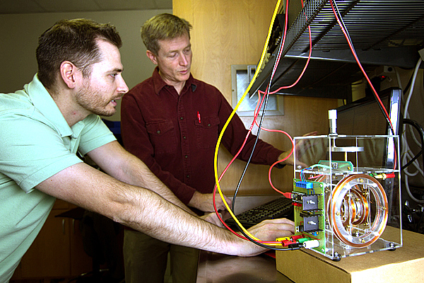 Two men, a student in light green and a teacher in brown, presumably, stand over a contraption (a clear plastic box with coils and wires inside) that appears to be connected to a computer resting on the counter top.