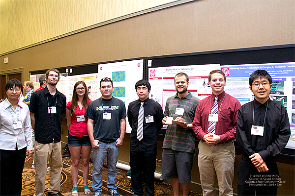 A group of eight (8) students stand, shoulder to shoulder, posed in a lobby of sorts, in front of several science presentations mounted on the wall behind them.