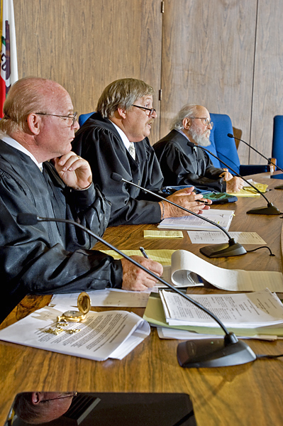 Three men in black robes sit on a panel with microphones pointed toward them. They sit in what appears to be a courtroom.