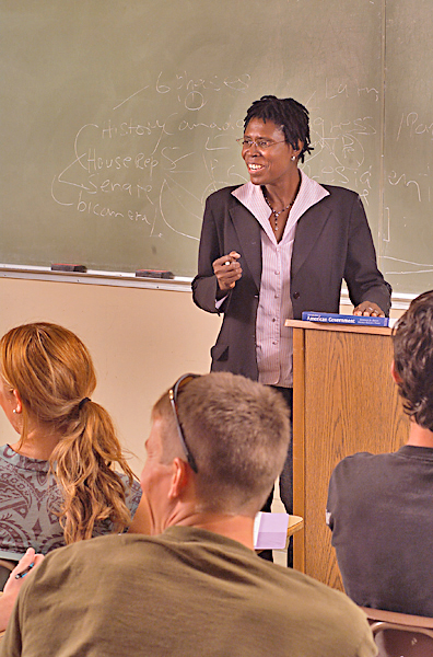 Female lecturer stands next to podium with one hand on an "American Government" text book and holding chalk in her other hand actively listens to an unseen person. Others in desks look towards unseen person.
