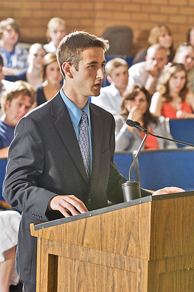 Man in business suit talks into microphones attached to a podium. Others in seats behind him watch and listen.