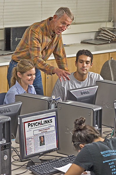 Older male leans over two seated students in a computer lab. He gestures toward the monitor of a female student while leaning on a male student's chair. "Psych-LInks" appears on the computer screen of another female student.