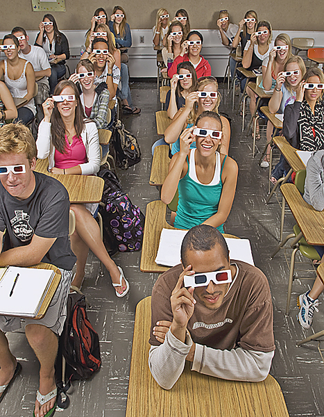 A classroom of students in rows of desks hold 3D glasses up to their eyes.