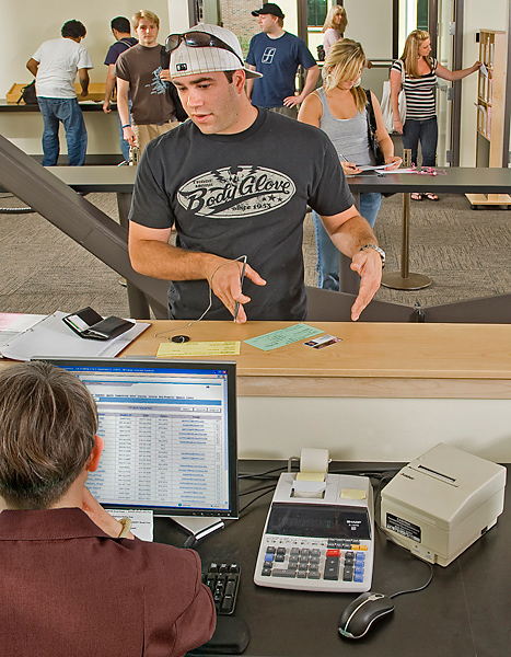 Man wears backwards ball cap standing at counter to fills out forms. His Wildcat ID, wallet, and binder sit on the counter. He is gesturing to the right, looking at a woman sitting behind the counter.