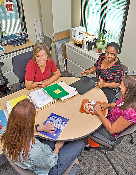 Four women sit at a round table in a corner office smiling with binders and booklets on the table in front of them.