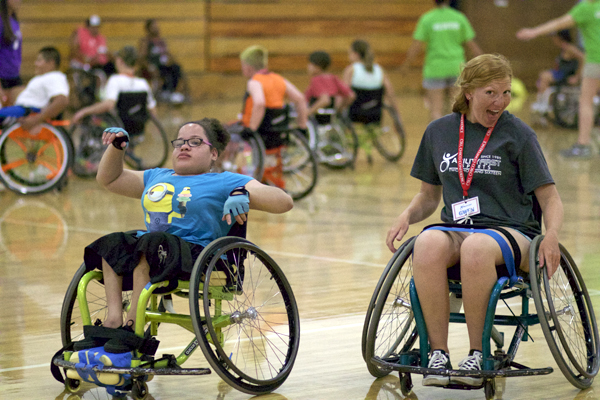 Two women in wheelchairs are in a gymnasium with several people in wheelchairs in the background.  Woman on left is wearing a turquoise "Minions" t-shirt and matching cut-off gloves.
