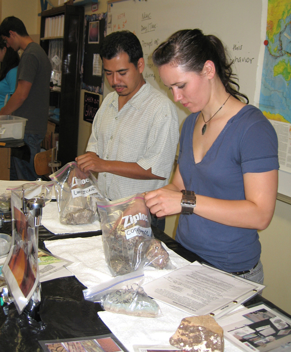 Male and female stand at lab table each looking down, removing rock specimen from Ziploc bag.