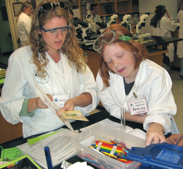Woman in lab coat holds small poster while girl in lab coat picks through colored pencils.