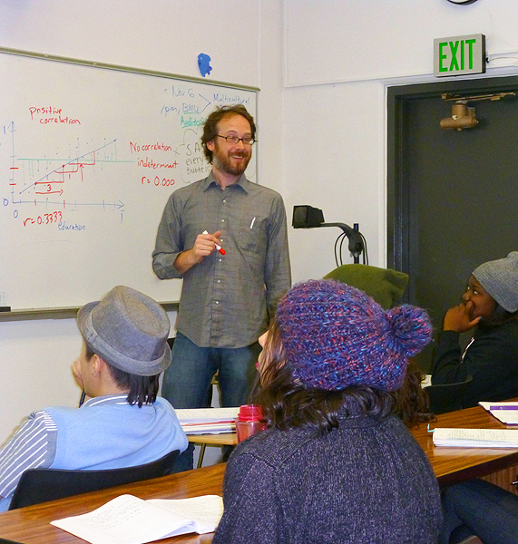 A bearded smiling man, holding a red dry-erase marker, stands between a group of students seated in a desk and a dry-erase board that has a graph and other writing.  The students in the shot all wear hats.  One individual wears a derby while the other three wear knit-caps.
