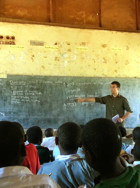 A man in an olive green dress shirt holds a booklet in his left hand while pointing to the word "vernacular" written on a chalkboard with a blurred right hand.  He stands in front of a group of dark-skinned young boys who all focus on him.  In the back of the group (image foreground) one boy leans to his left possibly to get a better view.