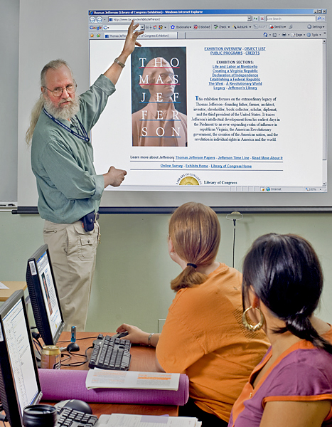 Bearded man with pony tail and glasses gestures to a projected image of what appears to be the course syllabus.