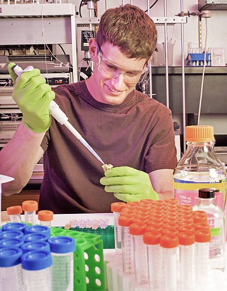 Man wearing goggles and green latex gloves uses a large syringe to inject a fluid into a test tube.
