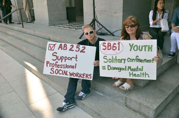 One man and one woman sit on a set of granite stairs, wearing sunglasses. Each holds a protest sign.  The man, dressed in dark colors holds a sign with a red check mark on it reading "AB 252: Support Professional Social Workers."  The sign the woman holds has "SB61" circled and X'd out. The sign reads: "Youth Solitary confinement=Damaged Mental Health."