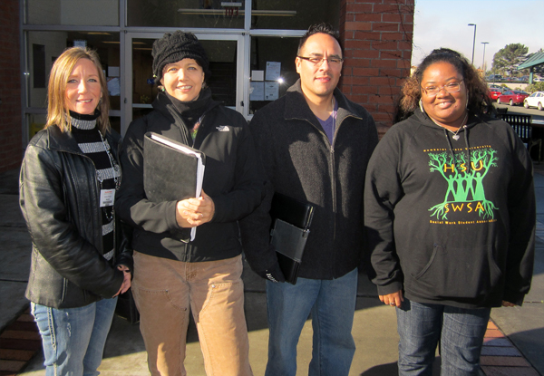 Four people, three women and one man, stand outside in front of a building entrance. All individuals wear black. The two in the middle hold binders.  