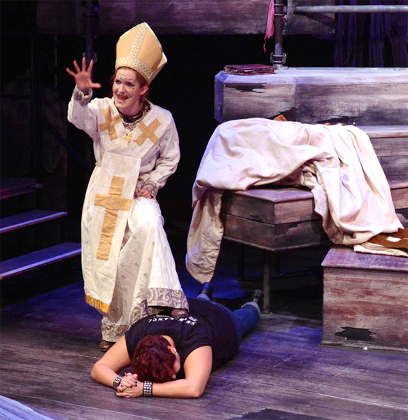 A woman with hand out stretched reaching towards the viewer, in exaggerated make-up wearing a white archbishop robe trimmed in gold with gold crosses and a gold headrest, stands with one foot on a dark-haired man wearing a black vest and blue jeans who lies face down with his hands clasped together in front of his head. Three coffins are stacked on the right. Stairs lead off to the left.