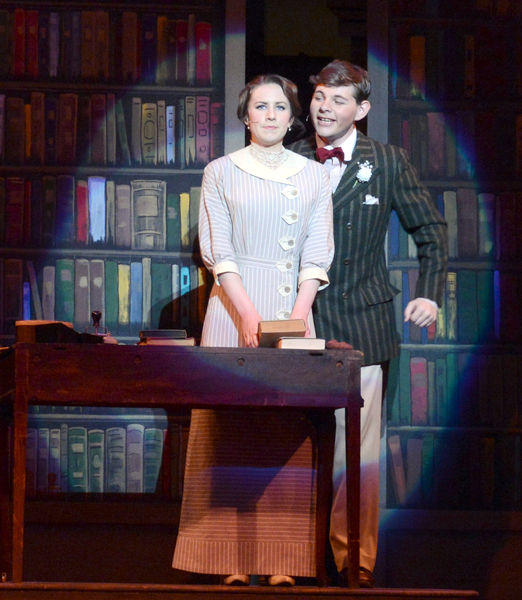 Man and woman, dressed in early 20th-century stand in a spotlight in behind a table in front of a bookcase backdrop. She stands in front of him, both hands holding a book just above the table, wearing a light-colored three quarter length sleeved dress with pinstripes, circular collar, and half-dollar sized buttons down the left side of the front. He wears a black pinstriped, double-breasted suit jacket, red bow tie, and light trousers, gazing at her grinning. Each wears a headset microphone.