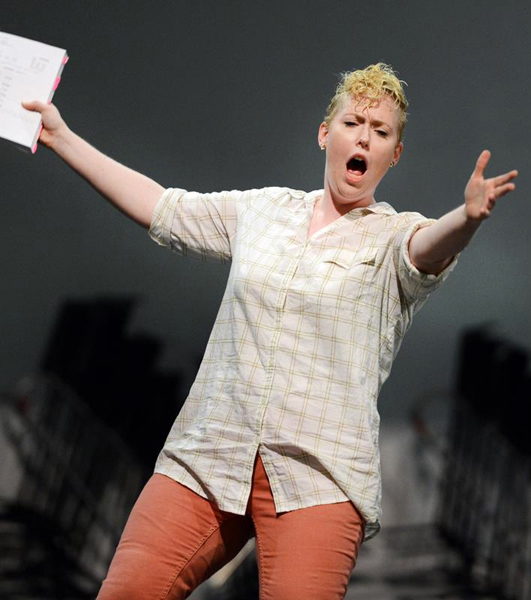 A woman, informally dressed in a grid patterned button down blouse with burnt orange pants, leans to the right with one arm outstretched towards the viewer, fingers spread, and the other arm stretched out to her side holding what appears to be a large script with pink tabs poking out. Her hair is blonde and curled in front and her mouth is agape as if in the middle of singing  a note.