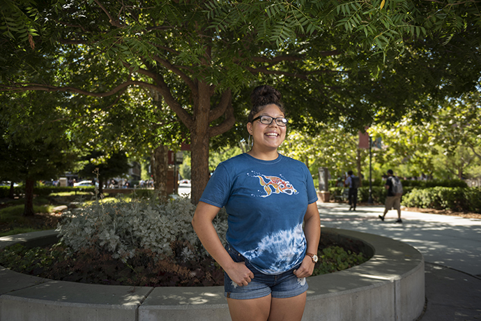 Student Jacklynn Rodriguez stands in front of a tree in a planter on the Chico State campus with a smile on her face as she reflects on her college journey.