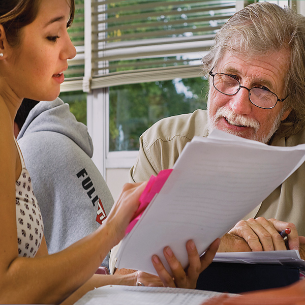 Older man (right) leans right peers at papers held by young woman (left) who points to page.