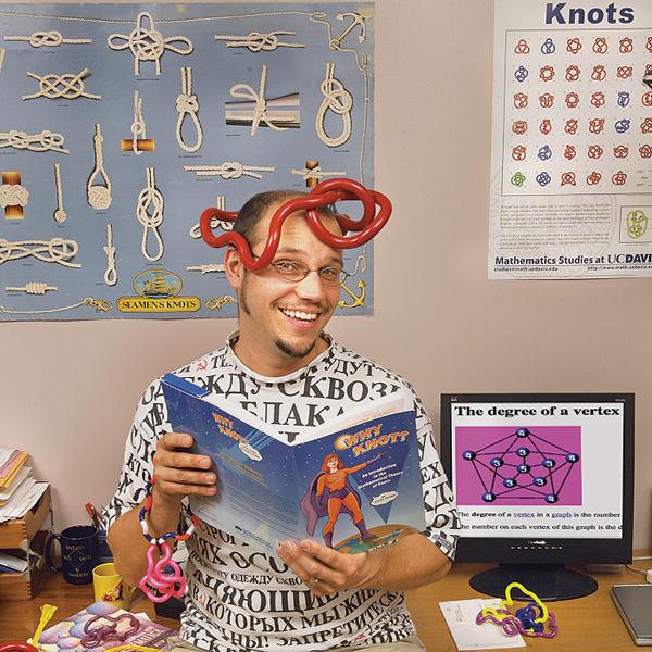 Man in glasses holds book (titled "Why Knot?") wearing a model of mathematical knots mirrored on a poster on the wall just to the side of the man.