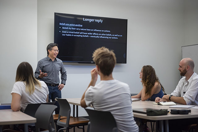 A man leads a discussion at the front of a classroom surrounded by four students who are facing him.