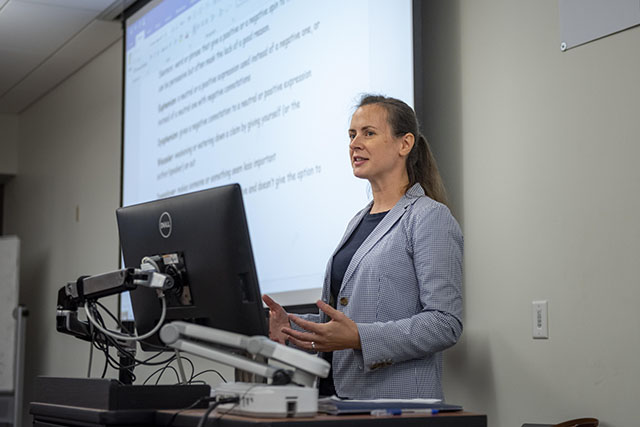 Woman in checkered blue blazer stands in front of a computer while words from her presentation is displayed behind her on a white screen as she gestures with her hands while she talks.