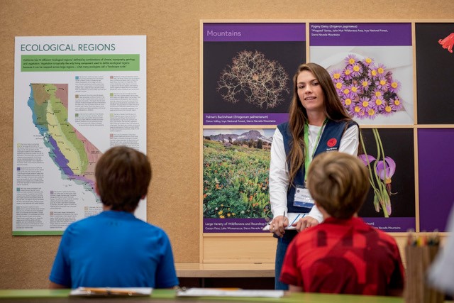 Science Ed female student (right) stands in front of two elementary school students who are facing her.