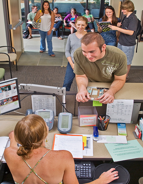 Man leans on counter to converse with woman sitting on the other side. One woman smiles waiting in line while two other people smile while perusing a book.