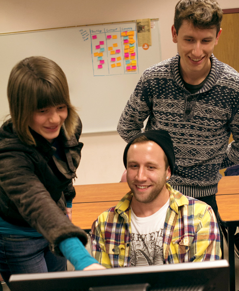 A man in a black stocking cap wearing a yellow plaid shirt sits in front of a computer.  A woman standing in a brown jacket leans in from the left to point at the computer monitor. Another man in an intricately patterned sweater smiles with his arms folded behind his back, stands behind the seated man, to the right of the picture.