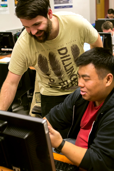 A dark-haired, bearded man in a pale yellow shirt with brown writing on it smiles as he leans over a seated man in a red shirt with a black jacket over it.  Both look and gesture towards a computer monitor in the lower left.