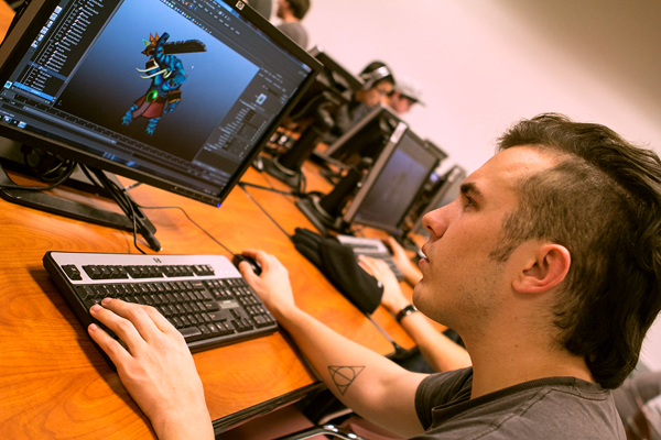 A tilted image shows a man sitting at a computer, facing the upper left, concentrates on an animated character on the computer screen of which he sits in front. His left hand is on the keyboard in the foreground while his right hand holds the mouse.  Several others sit just beyond him in a computer lab.