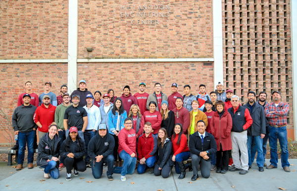 A group of students and professors smile and pose (standing and kneeling) in front of a brick wall (west side of Langdon Hall) on the CSU, Chico campus.