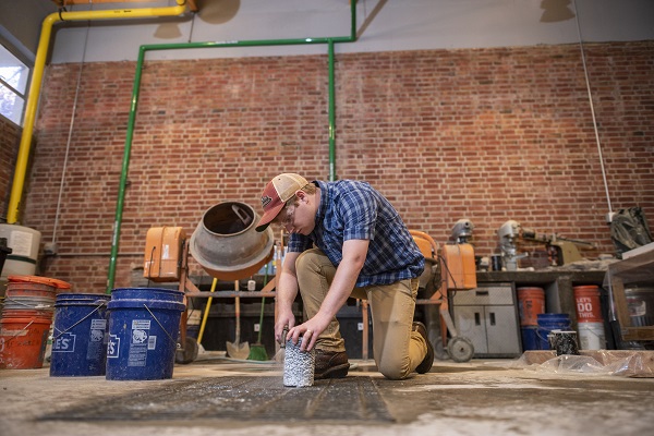 A man with a baseball cap kneels down on one knee while shaving a concrete block.